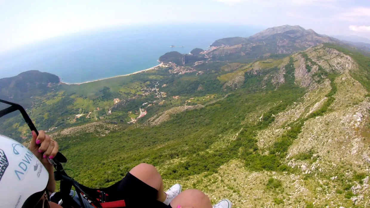 Paragliding above green coastal slopes with a bay and beach visible ahead.