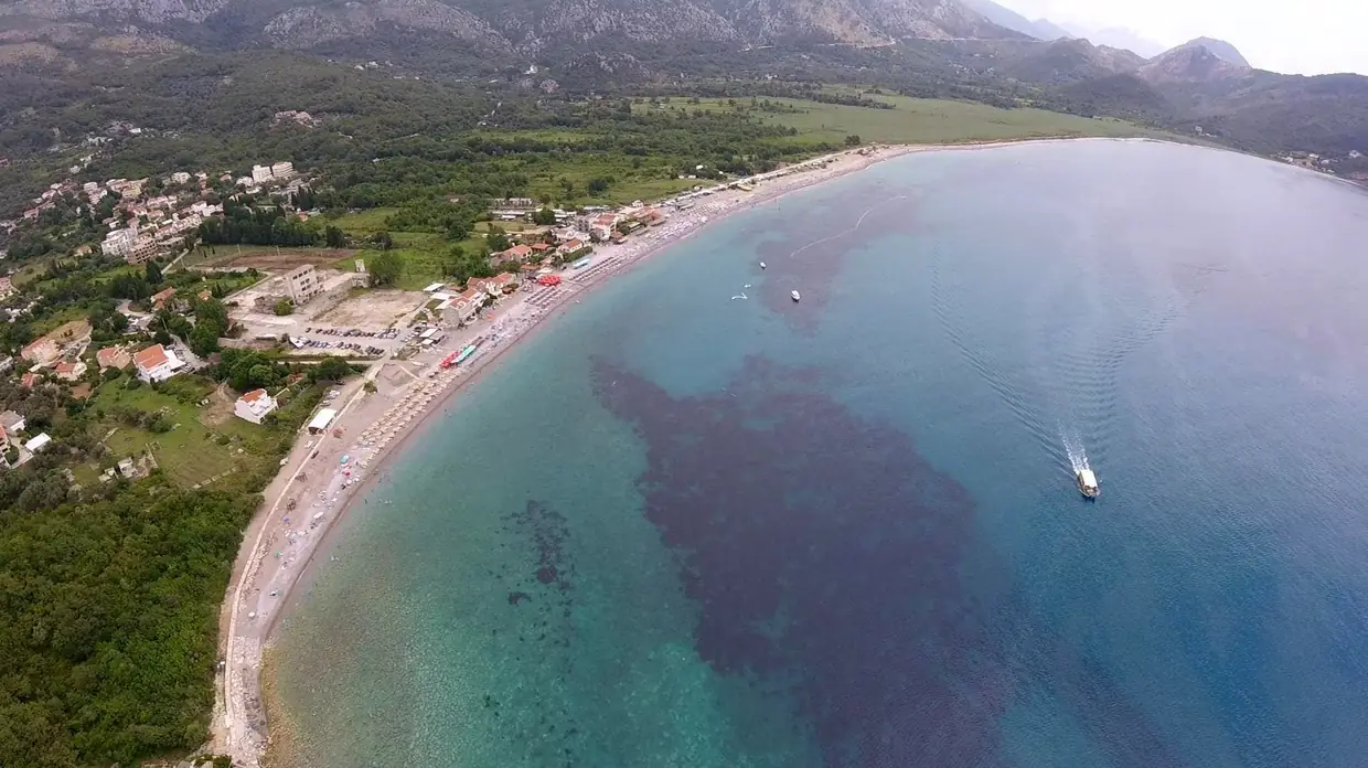 Aerial view of Buljarica bay with beach, coastal settlement, and mountains behind.